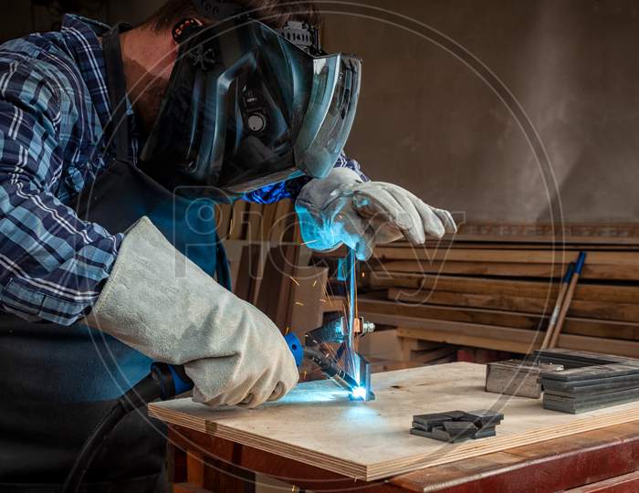 A Young  Man Welder In  Uniform, Welding Mask And Welders Leathers, Weld  Metal  With A Arc Welding Machine  In Workshop, Blue And Orange  Sparks Fly To The Sides