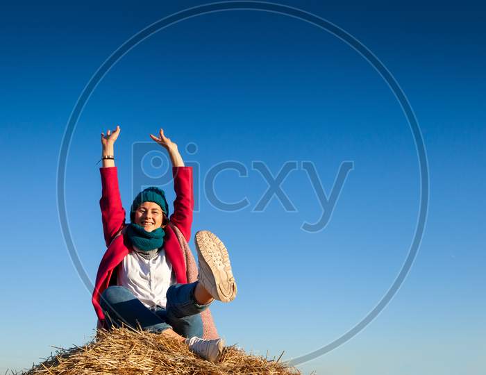 Portrait Female Student On Field Background. Beautiful Modern  Young Woman Wearing Pink Knitting Hat  Smile, Look At The Camera And Enjoy Autumn Sunny Day And Sitting On Haystick.