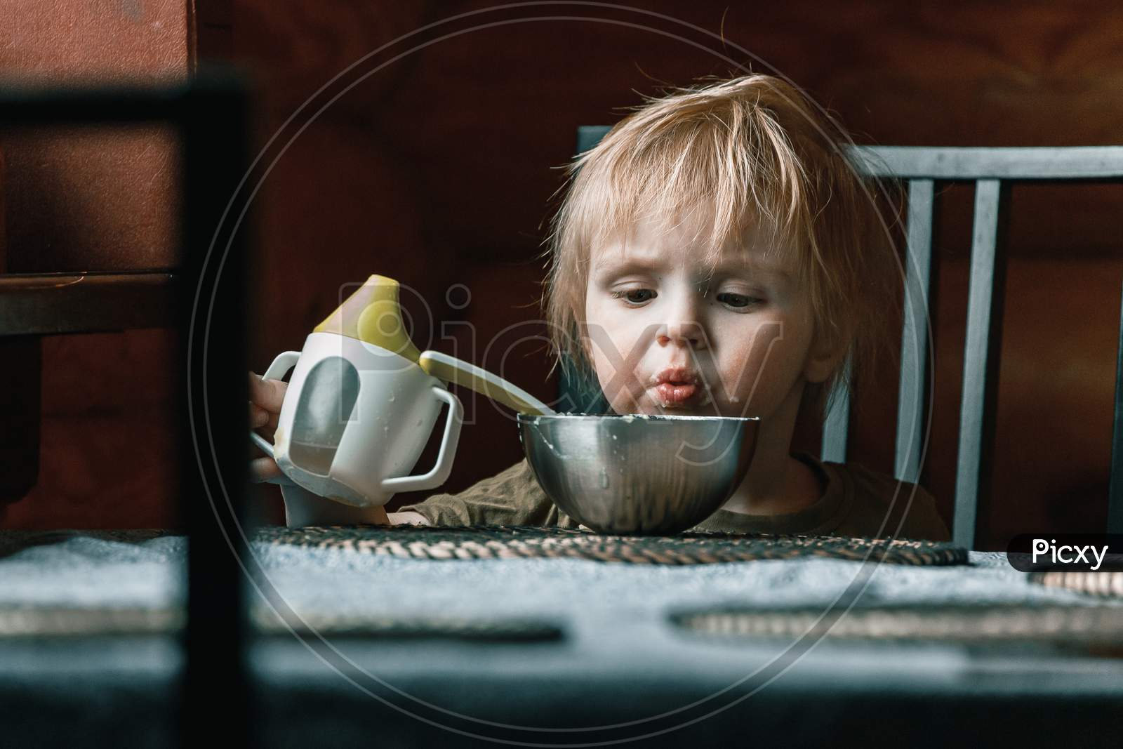 A Small Child Of Two Years Old In The Kitchen Eats Mashed Potatoes And Drinks Water From A Baby Sippy Cup At The Dining Table In The Kitchen