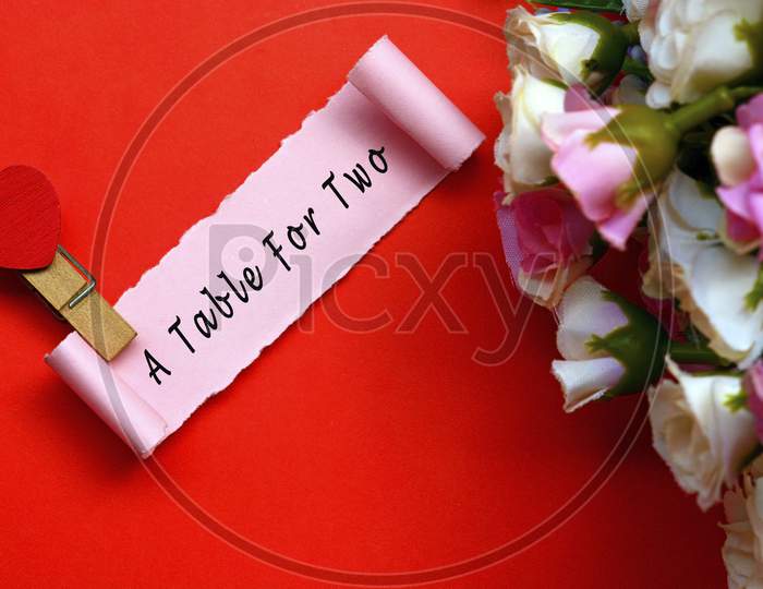 A Table For Two Label On Torn Paper With Flowers And Heart Shape Clothespin On Red Background. Valentine'S Day Concept