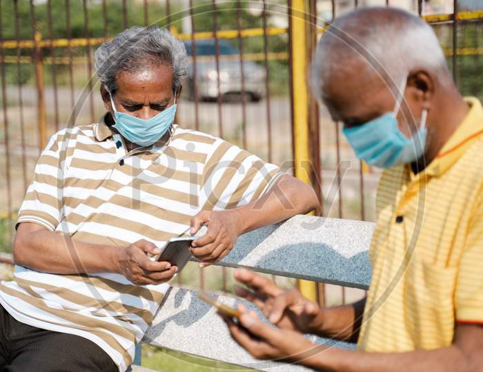 Sideview Of Two Senior Men With Medical Face Mask Using Mobile Phone While Sitting At Park By Maintaining Social Distance Concept Of Coronavirus Covid-19 Pandemic New Normal And Safety Measures.