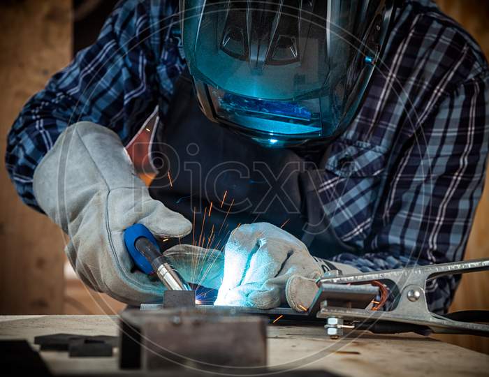 Close Up Of A Young  Man Welder In  Uniform, Welding Mask And Welders Leathers, Weld  Metal  With A Arc Welding Machine  In Workshop, Blue And Orange  Sparks Fly To The Sides