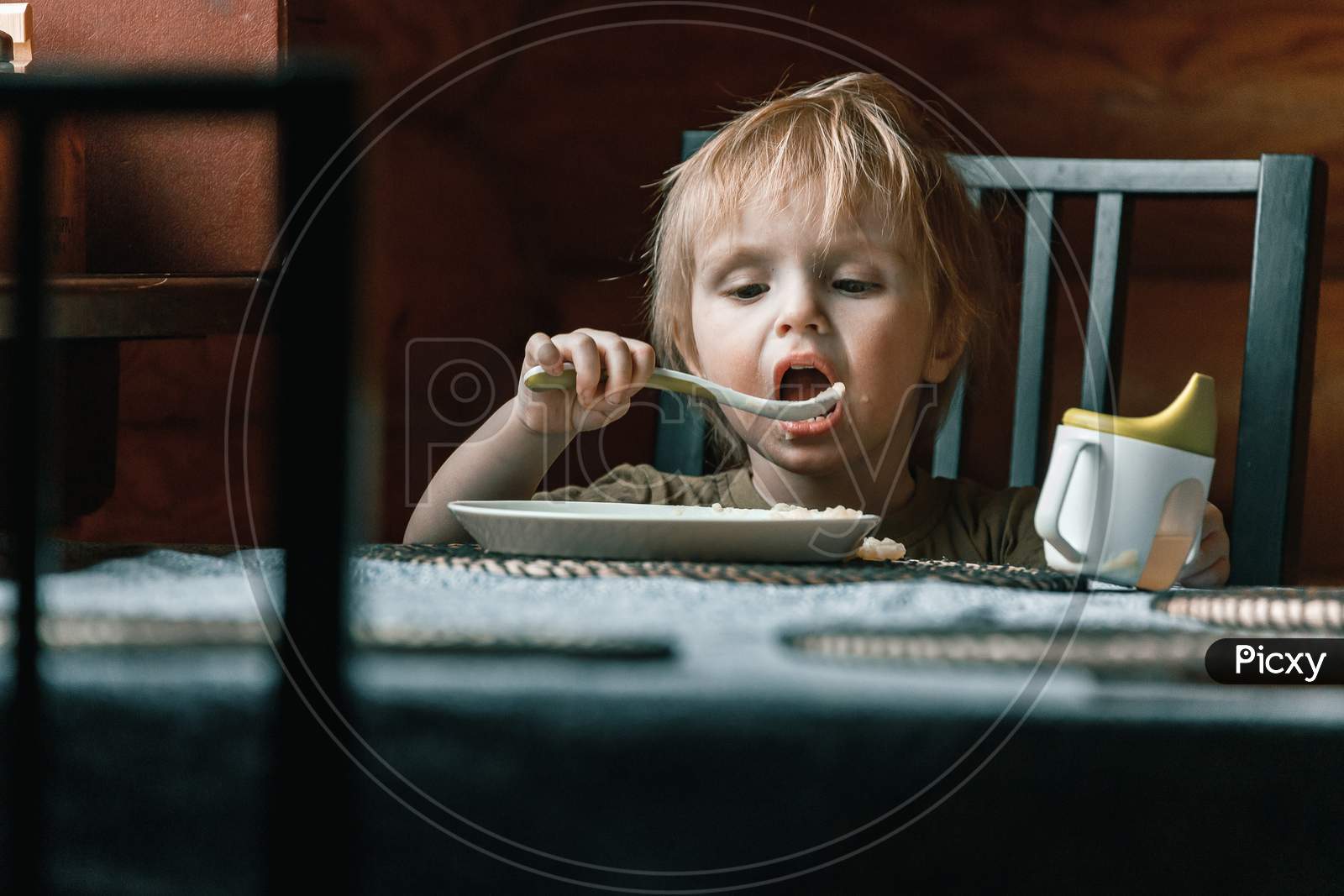 Close-Up Portrait Of A Little Blond Boy Of Two Years Old Who Eats With A Spoon Of Mashed Potatoes On His Own And Looks Pensively Ahead At Home