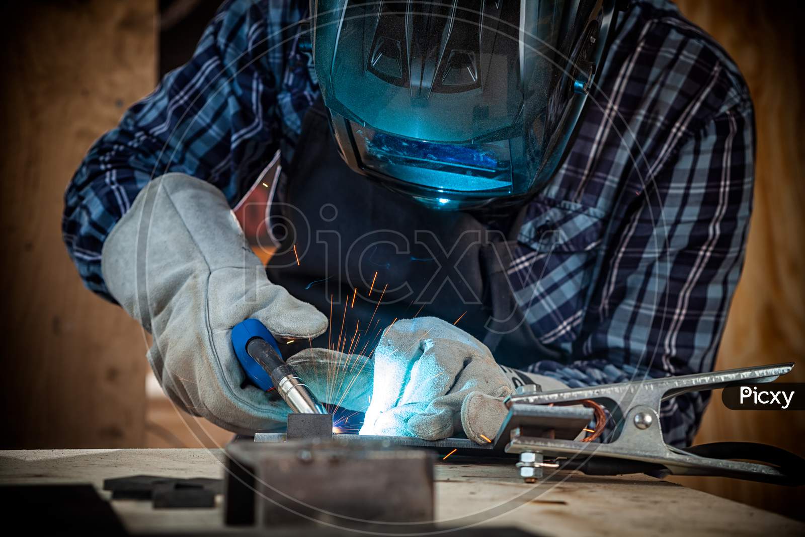 Close Up Of A Young  Man Welder In  Uniform, Welding Mask And Welders Leathers, Weld  Metal  With A Arc Welding Machine  In Workshop, Blue And Orange  Sparks Fly To The Sides