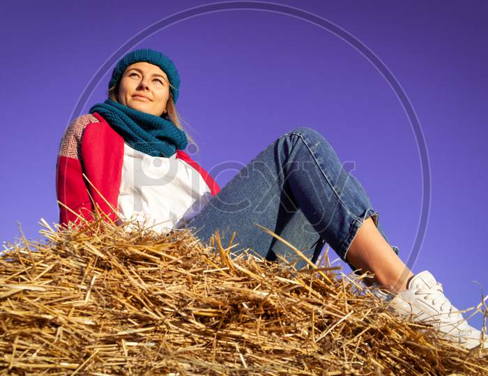 Fashion Lifestyle Portrait Of Young Trendy Woman Dressed In Pink Coat, Pink Knitted Hat  Posing, Laughing And Smiling On A Haystack Around Blue Sky.  Portrait Of Joyful Woman