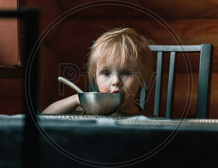 Close-Up Portrait Of A Little Blond Boy Of Two Years Old Who Eats With A Spoon Of Mashed Potatoes On His Own And Looks Pensively Ahead At Home