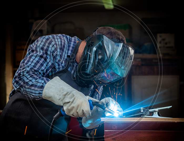 A Strong Man Welder In A Black T-Shirt, In A Welding Mask And Welders Leathers Weld Metal Welding Machine In The Workshop, In The Sides Fly The Blue Sparks