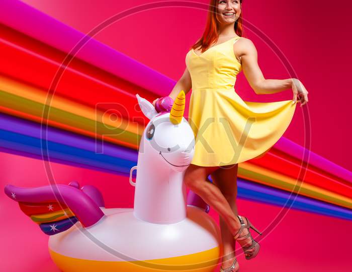 Concept Summer Mood, Relaxation And Beauty.Modern Portrait Of A Young Woman In A Yellow Dress Resting On An Inflatable Unicorn Mattress On An Isolated Pink Background With Rainbow
