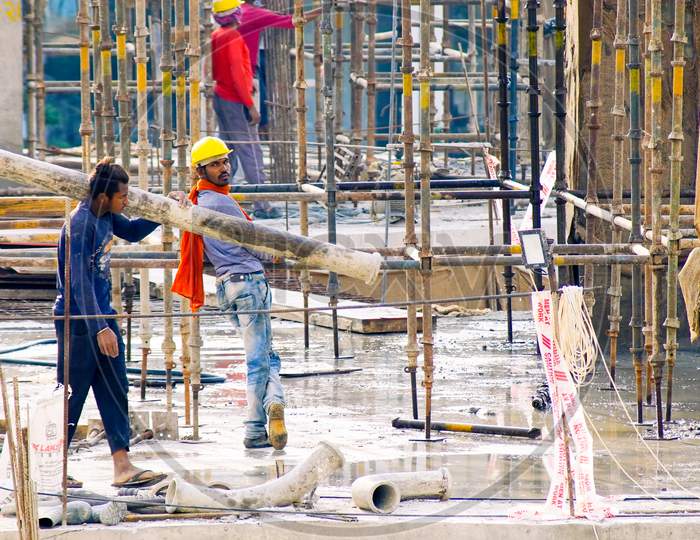 Construction Site In India At Dusk With Workers, People, Labor, Engineers In Colorful Clothes Among Rebar Wood Concrete Pillars Working And Carrying A Pipe While Another Looks On