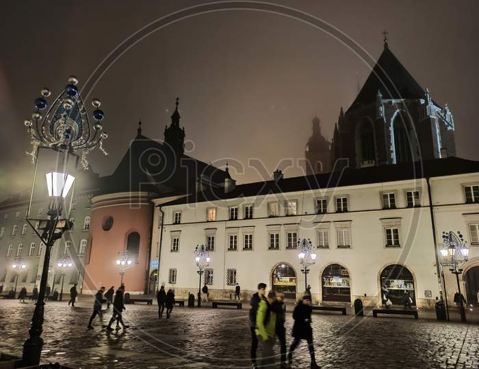 Krakow, Poland - November 08, 2019: Wide Angle View Of The Little Market Square, Maly Rynek, In Old Town Of Cracow And Long Exposure Shot Of A People Walking At Night During Foggy Weather In Winter