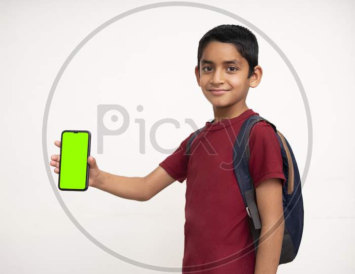 Young Indian Kid Holding A Phone In His Hand With A Green Screen, School Bag In His Back And Wearing Red T-Shirt Standing On White Isolated Background.
