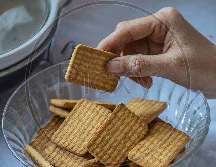 Stock Photo Of A Women Taking Wheat Biscuit From A Bowl Of Biscuits , Kept On Table On White Background. Focus On Biscuit.