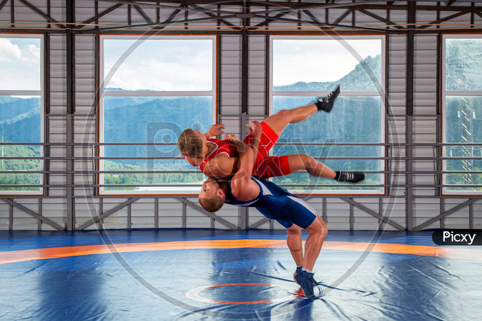 Two Young Men In Blue And Red Wrestling Tights Are Wrestlng And Making A Suplex Wrestling On A  Blue Wrestling Carpet In The Gym On The Background Of Mountains. The Concept Of Male Wrestling And Resistance