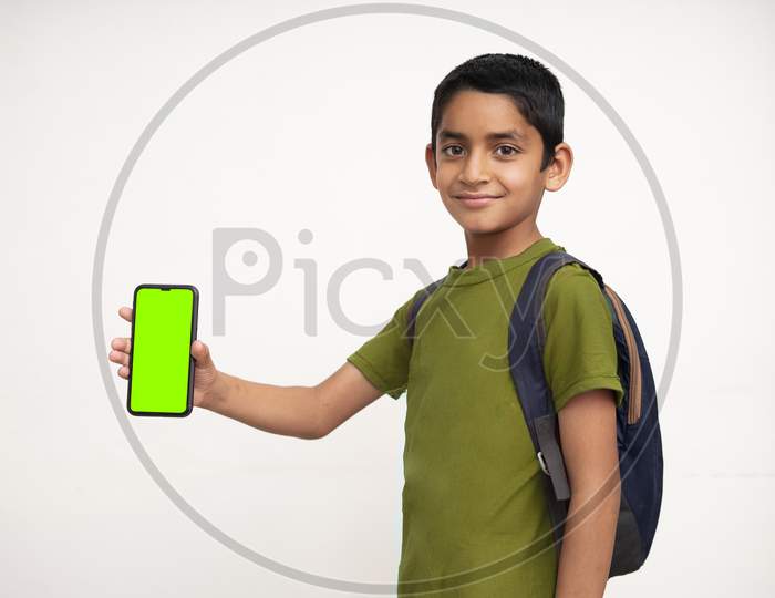 Young Indian Kid Holding A Phone In His Hand With A Green Screen, School Bag In His Back And Wearing Green T-Shirt Standing On White Isolated Background.