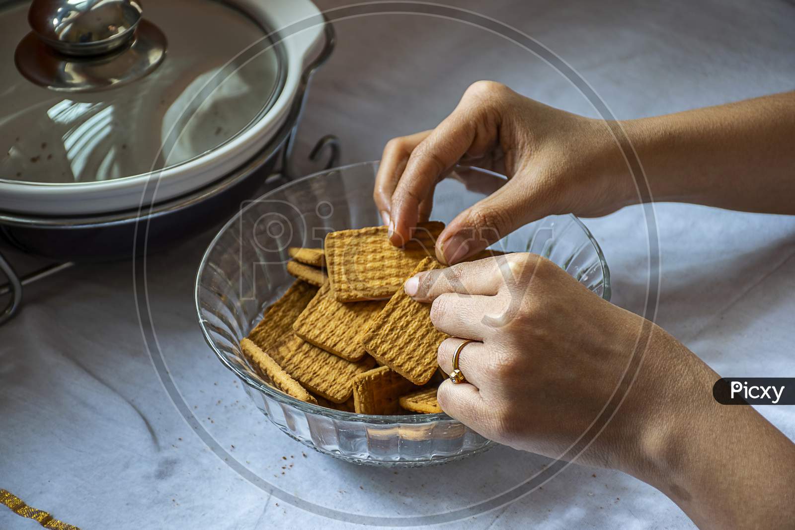 Stock Photo Of A Women Taking Wheat Biscuit From A Bowl Of Biscuits , Kept On Table On White Background. Focus On Biscuit.