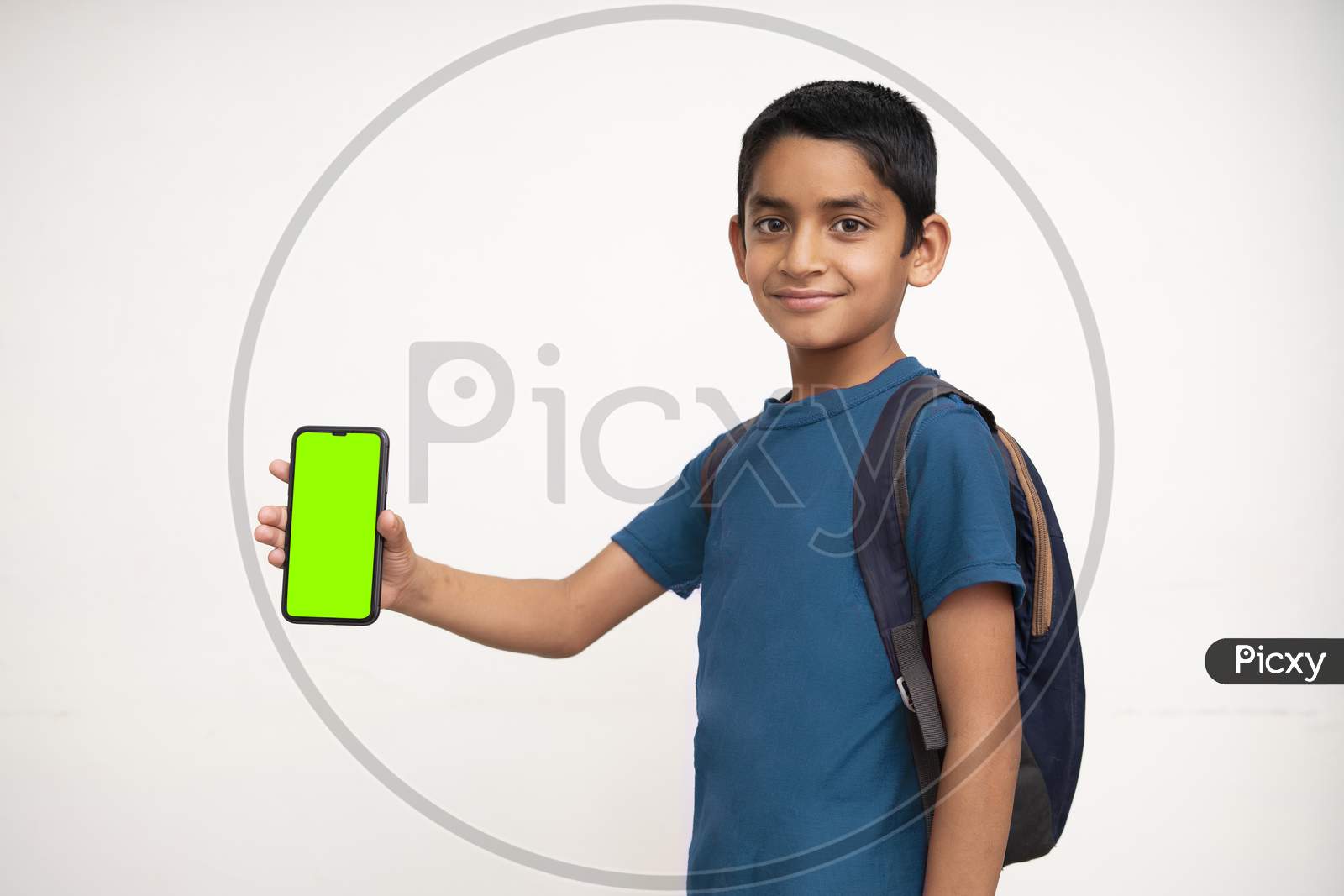 Young Indian Kid Holding A Phone In His Hand With A Green Screen, School Bag In His Back And Wearing Blue T-Shirt Standing On White Isolated Background.