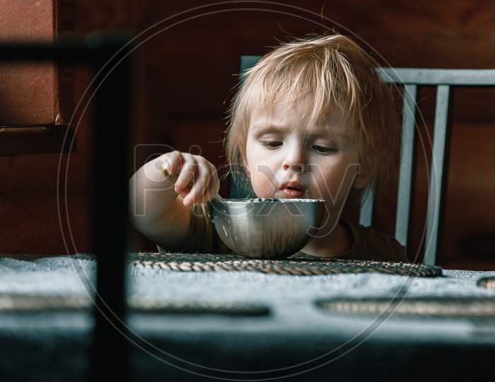 A Toddler Boy On A Chair Holds A Plastic Spoon And Eats His Lunch. Feeding Process. An Infant Child Learns To Eat. Baby Food, Family, Child, Food