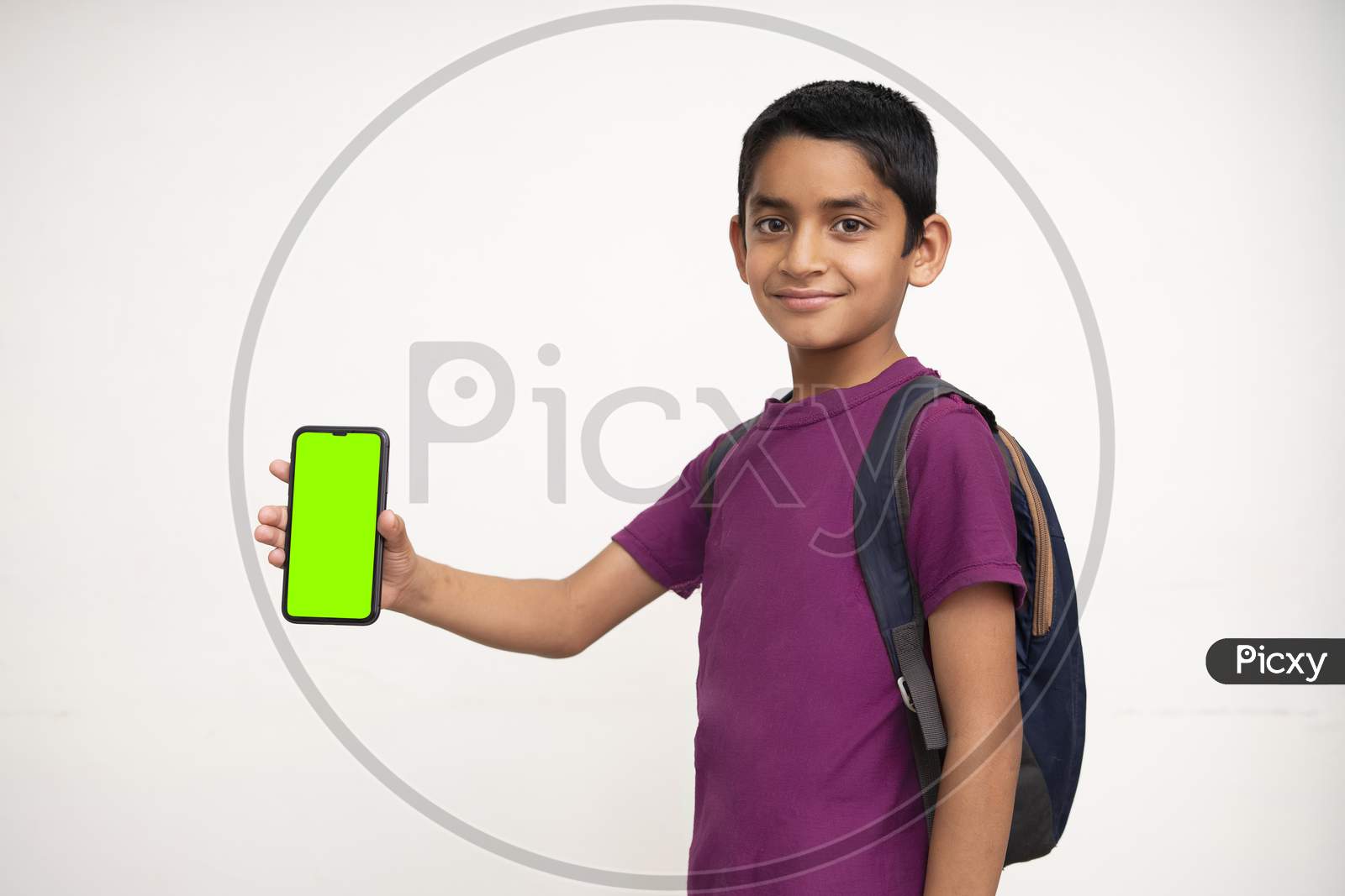 Young Indian Kid Holding A Phone In His Hand With A Green Screen, School Bag In His Back And Wearing Purple T-Shirt Standing On White Isolated Background.