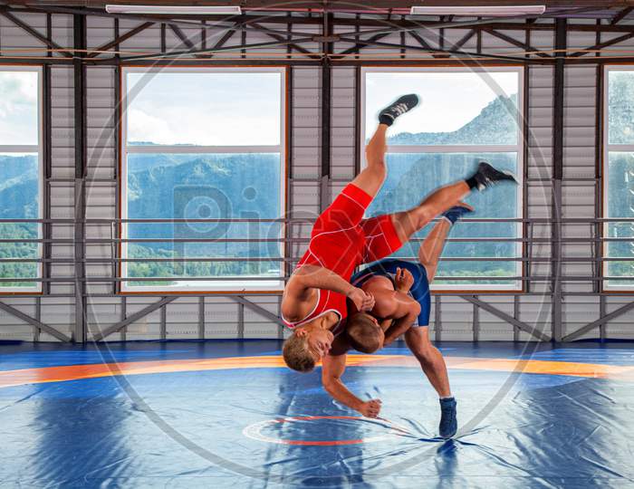 Two Young Men In Blue And Red Wrestling Tights Are Wrestlng And Making Shot Through The Thigh On A  Blue Wrestling Carpet In The Gym On The Background Of Mountains. The Concept Of Male Wrestling And Resistance