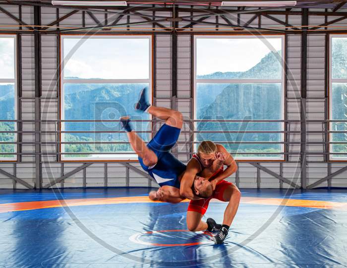 Greco-Roman Wrestling Training, Grappling. Two Greco-Roman  Wrestlers In Red And Blue Uniform Making A Thigh Throw  On A Wrestling Carpet In The Gym.The Concept Of Male Wrestling And Resistance