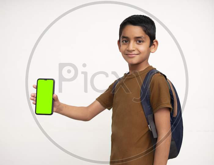 Young Indian Kid Holding A Phone In His Hand With A Green Screen, School Bag In His Back And Wearing Brown T-Shirt Standing On White Isolated Background.