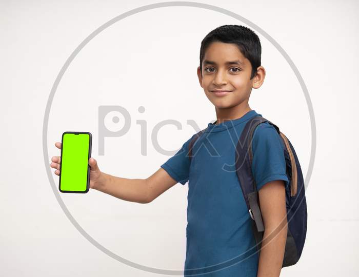 Young Indian Kid Holding A Phone In His Hand With A Green Screen, School Bag In His Back And Wearing Blue T-Shirt Standing On White Isolated Background.