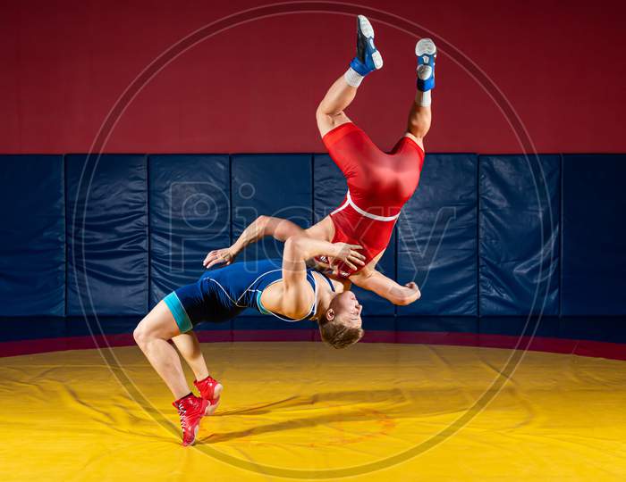 Two  Strong Men In Blue And Red Wrestling Tights Are Wrestlng And Making A Suplex Wrestling On A Yellow Wrestling Carpet In The Gym. Wrestlers Doing Grapple.