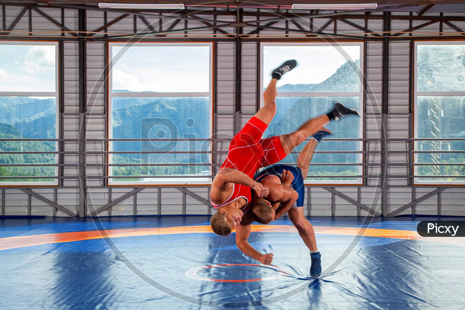 Two Young Men In Blue And Red Wrestling Tights Are Wrestlng And Making Shot Through The Thigh On A  Blue Wrestling Carpet In The Gym On The Background Of Mountains. The Concept Of Male Wrestling And Resistance