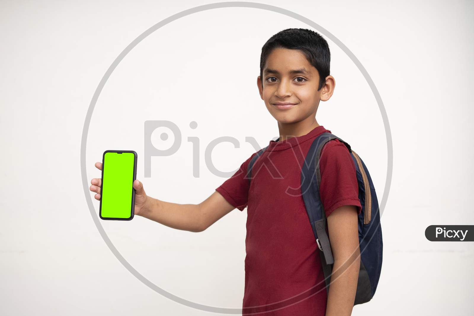 Young Indian Kid Holding A Phone In His Hand With A Green Screen, School Bag In His Back And Wearing Red T-Shirt Standing On White Isolated Background.
