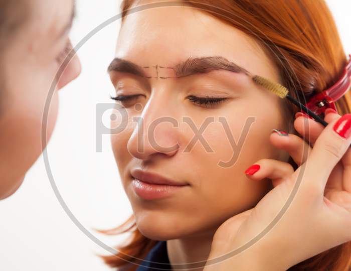 The Procedure For Eyebrow Correction In The Beauty Salon. The Make-Up Artist Penciled An Ideal Eyebrow Shape On A Young Red-Haired Woman And Combs Them With A Special Brush On A White Background