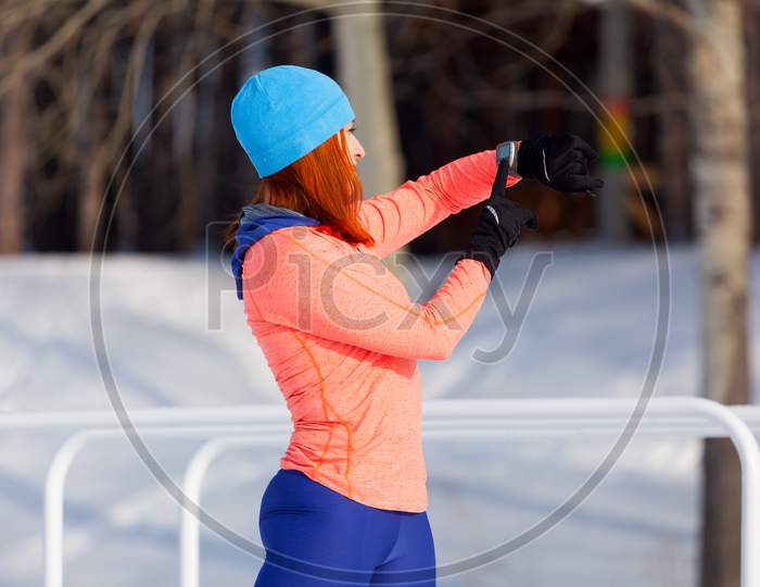A Young Woman In A Bright Blue Hat, Orange Sweater And Elk Smiles, Prepares For Training And Looks At The Sports Watch Before Running On The Playground On A Bright Winter Day