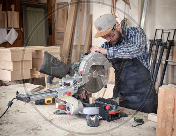 Experienced Carpenter In Work Clothes And Small Buiness Owner Working In Woodwork Workshop, Using A Circular Saw To Cut Through A Wooden Plank, On The Table Is A Hammer And Many Tools