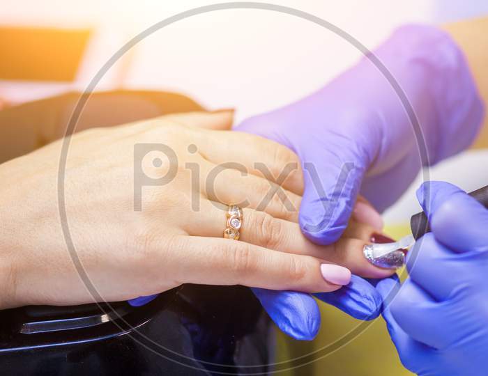 Close-Up Of A Manicure Master In Sterile Blue Gloves Paints A Girl'S Nails With A Protective Nail Polish On A White Manicure Table