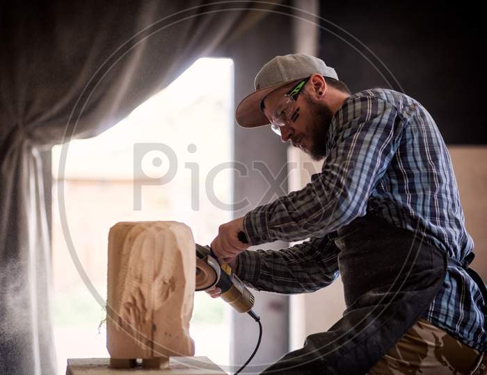 Сarpenter, Builder In Work Clothes Saw To Cut Out Sculpture From Wooden A Man'S Head, Using An Angle Grinder  In The Workshop, Around A Lot Of Tools,Wooden,Furniture For Work