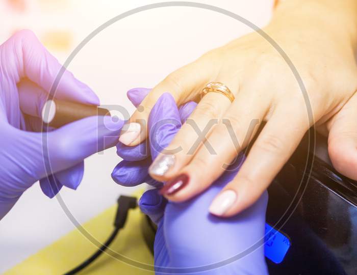 Close-Up Manicure Master With Sterile Blue Gloves Paints Nails With Protective Varnish To A Woman With Multi-Colored Nails On A White Table For Manicure