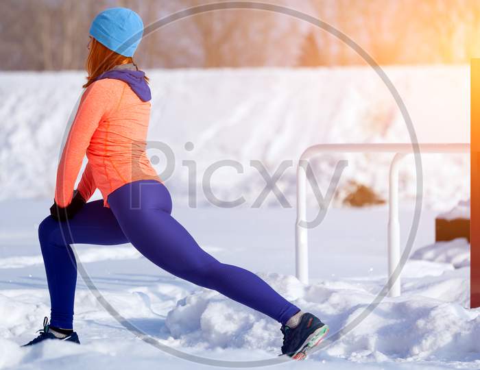 Young Woman In A Bright Blue Hat, Orange Sweater And Elk  Stretches Before Running On A Sports Field On A Bright Winter Day