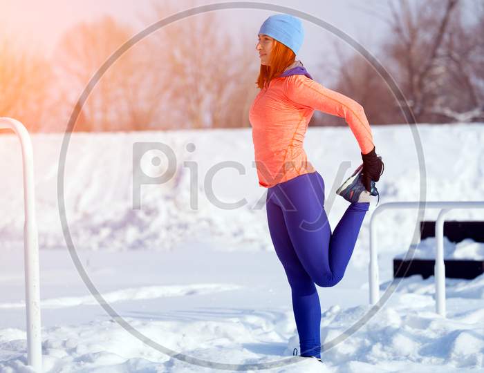 Young Woman In A Bright Blue Hat, Orange Sweater And Elk Smiles And Stretches Before Running On A Sports Field On A Bright Winter Day