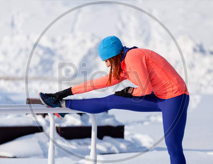 Young Woman In A Bright Blue Hat, Orange Sweater And Elk  Doing Stretching Before Running On A Sports Field On A Bright Winter Day