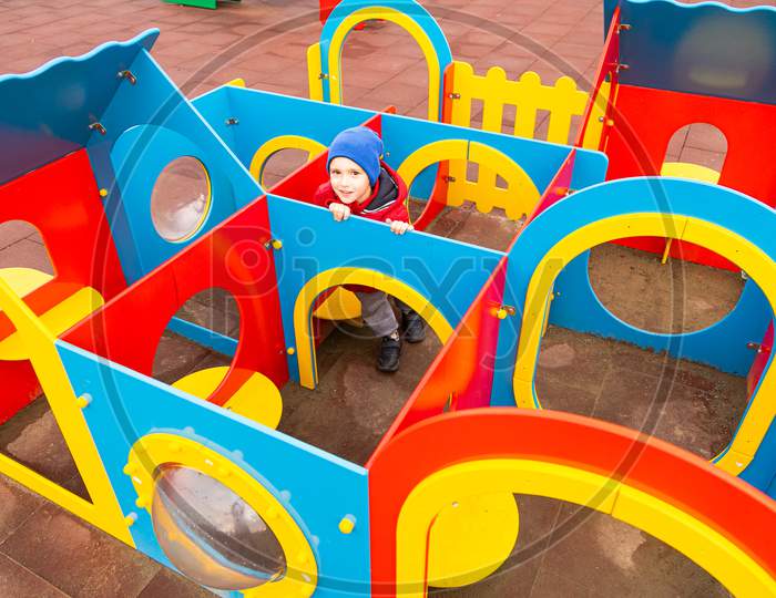 Little Cheerful Boy In A Warm Jacket, Hat Smiles And Plays In A Colorful Children'S Labyrinth On A Playground In A City Park. The Concept Of Kindergarten Fun And City Fun, Kindergarten Preschool Walks