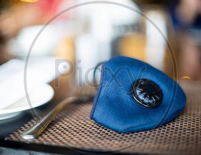 Blue Cloth Mask With Filter Placed On A Restuarant Table With Cutlery Knife And Fork Shot With An Out Of Focus Background