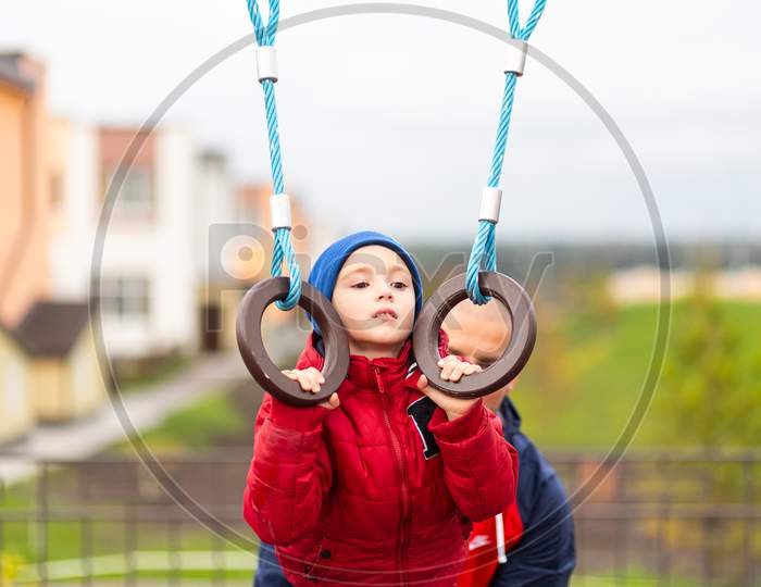 Little Funny Boy In A Warm Red Jacket And Hat Is Pulled Up On The Rings With The Help Of Dad'S Playground In The City Park. The Concept Of Children'S Entertainment, Communication And Learning Dad And Son. Father'S Day
