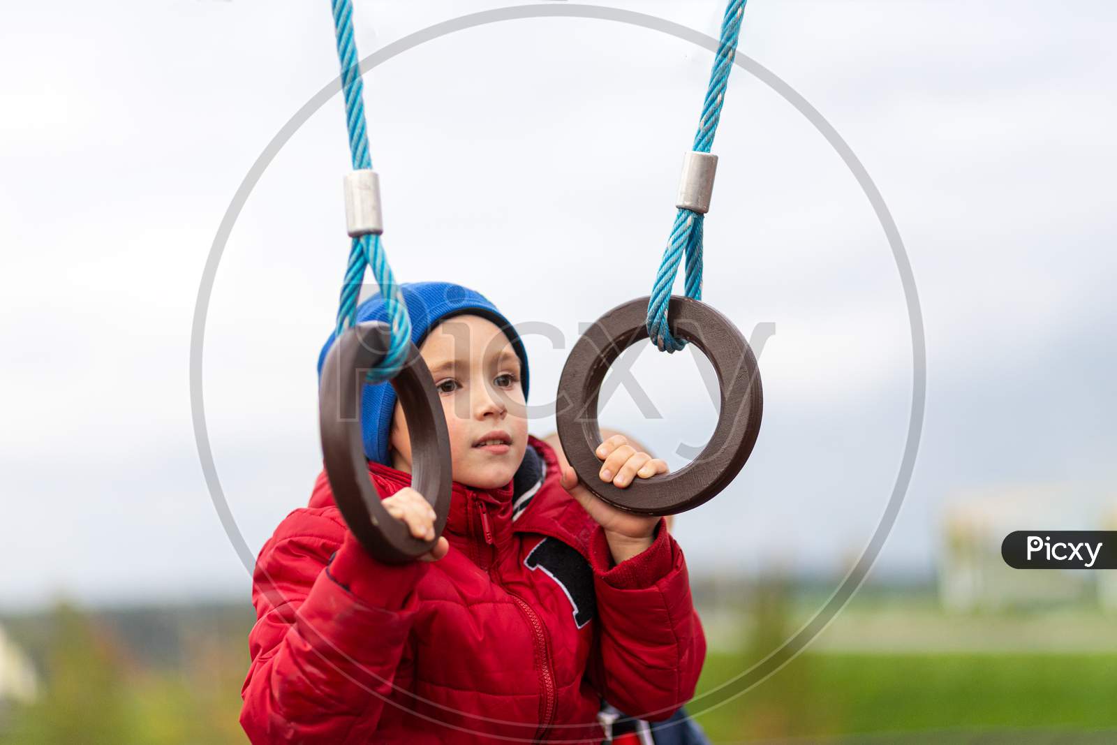 Little Funny Boy In A Warm Red Jacket And Hat Is Pulled Up On The Rings With The Help Of Dad'S Playground In The City Park. The Concept Of Children'S Entertainment, Communication And Learning Dad And Son. Father'S Day