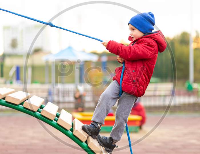 A Little Funny Boy In A Warm Red Jacket And Hat Climbs A Wooden Slide Using A Rope At A Playground In A City Park. Children'S Entertainment Concept