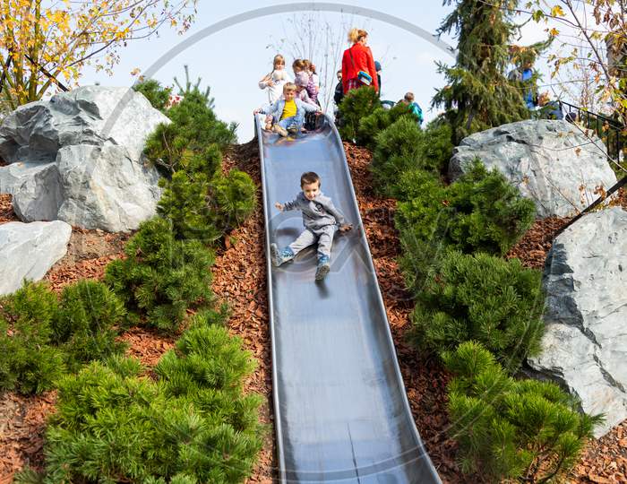 Little Cheerful Boy In A Tracksuit Smiles And Rides Out In A High Metal Slide On A Playground In A City Park. The Concept Of Kindergarten Fun And City Fun, Kindergarten Preschool Walks