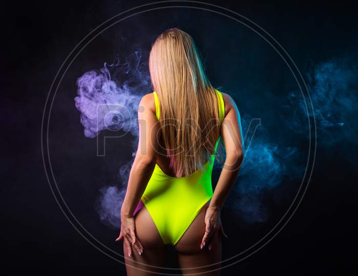 Young Dark-Haired Woman In Yellow Swimsuit Posing Against A Background Of Purple  And   Blue Smoke From A Vape On A Black Isolated Background