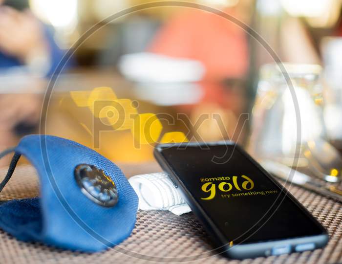 Blue Cloth Mask With Mobile Phone At Side With A Zomato Gold Logo In Restaurant Showing The Popularity Fo Safe Eating Out And Ordering In As Coronavirus Cases Increase