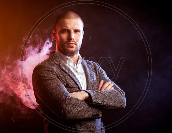 Young Handsome Man Manager In White Shirt And  Gray Suit  Stands With His Arms Crossed And Looks At The Camera Against Red Smoke From A Vape  On Black Isolated