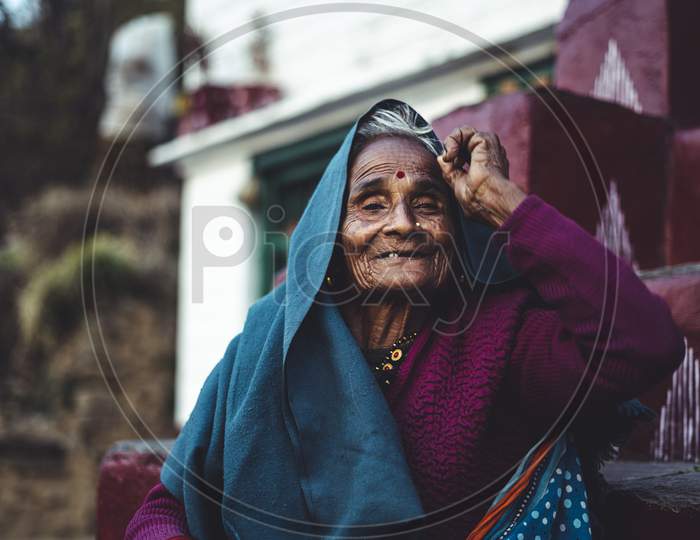 Almora, Uttrakhand - March 2 2021:- Old Aged Woman Sitting With The Support Of The Wall In The Sunset Wearing Indian Traditional Dress And Ancient Jeweleries