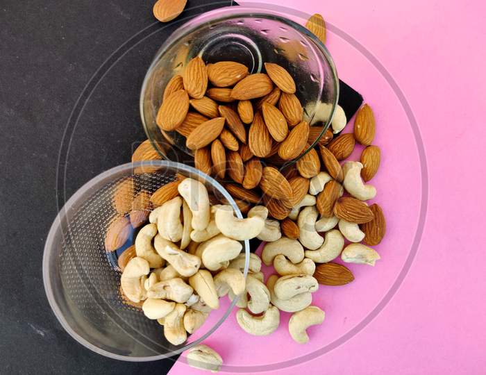 Top View Of Cashew Nuts And Almonds Split From Glass Bowl On Pink And Black Background
