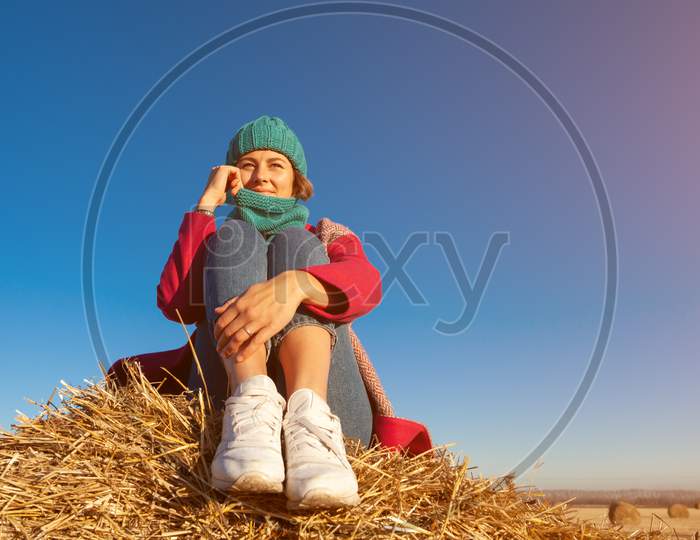 Young Attractive Lady In Blue Knitting Hat And Pink Coat Sitting On Haystack. Smiling Beautiful Hipster Happy Woman On Field, Wearing Stylish Clothes. Autumn Trend, Urban Stydent Style.